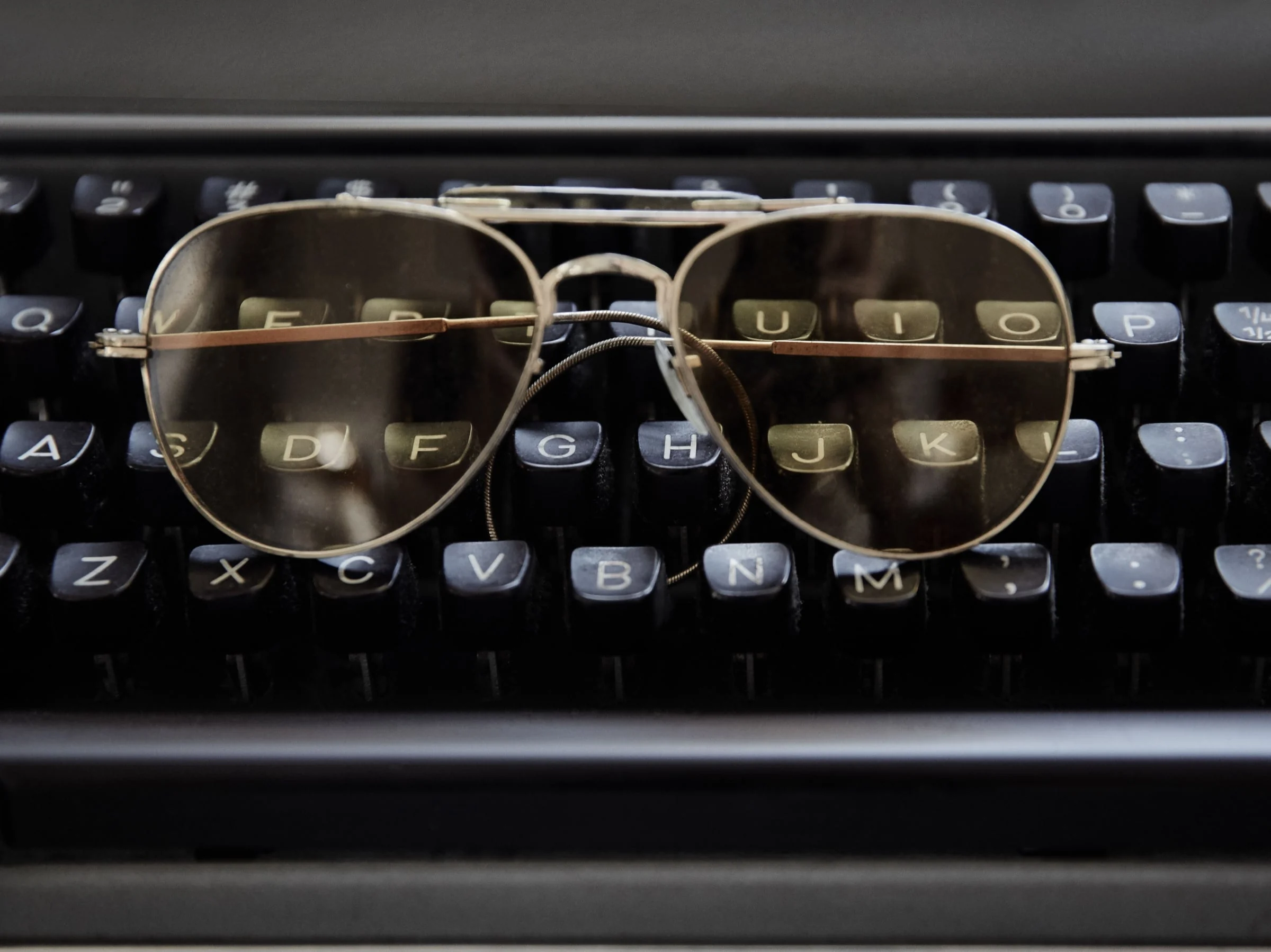 Gold wire aviator sunglasses with brown lenses resting on the black keys of a vintage typewriter, for Jeff Rudes.