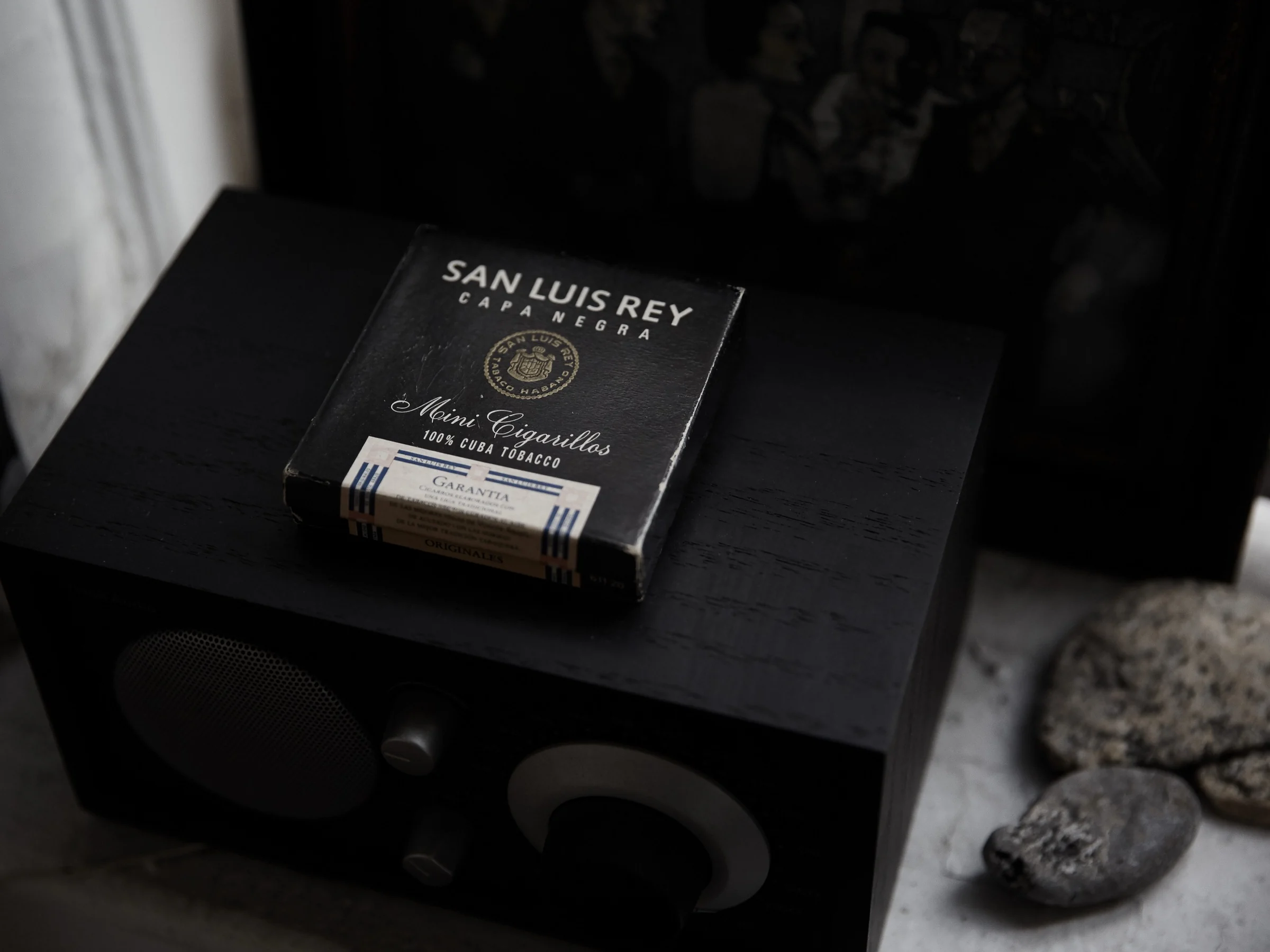 Box of San Luis Rey Capa Negra mini cigarillos on a black wooden table radio beside grey stones on a windowsill.