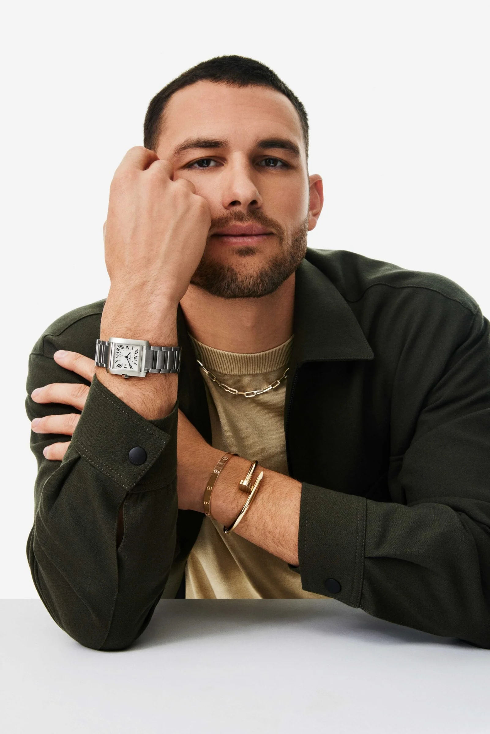 Man in olive jacket resting his head on his fist, wearing a steel Tank watch, Love and Juste un Clou bracelets, for Cartier.