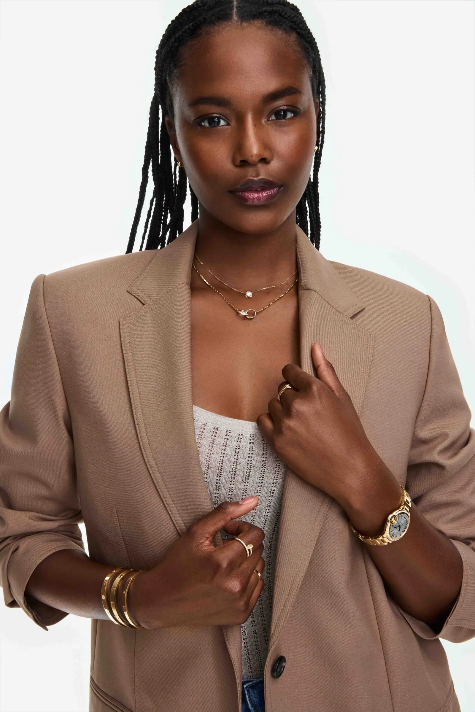 Model in a camel blazer and knit top wearing gold necklaces, rings, bangles and a gold watch on white, for Cartier.