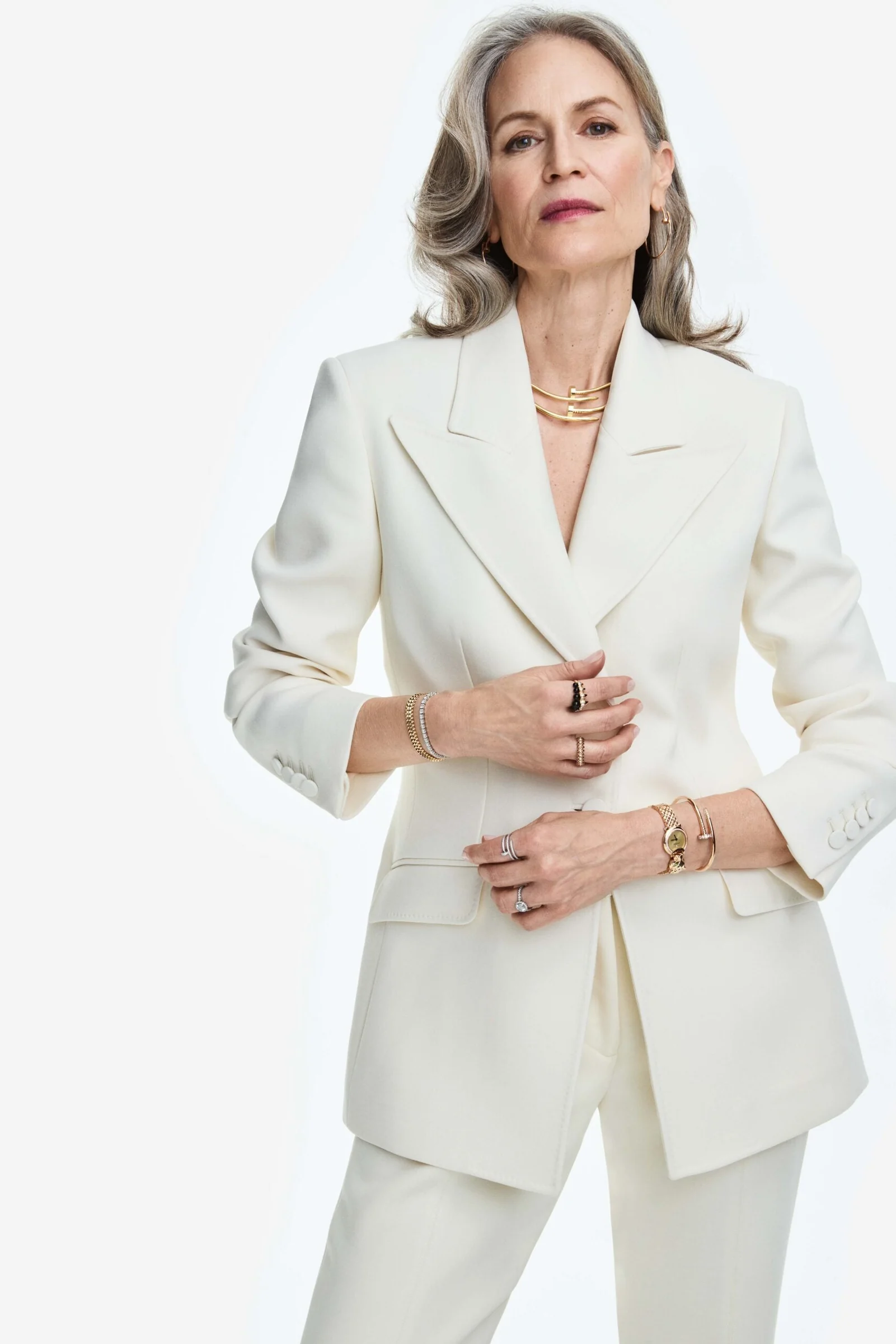 Grey-haired woman in an ivory double-breasted suit wearing gold necklaces, bracelets and rings on white, for Cartier.