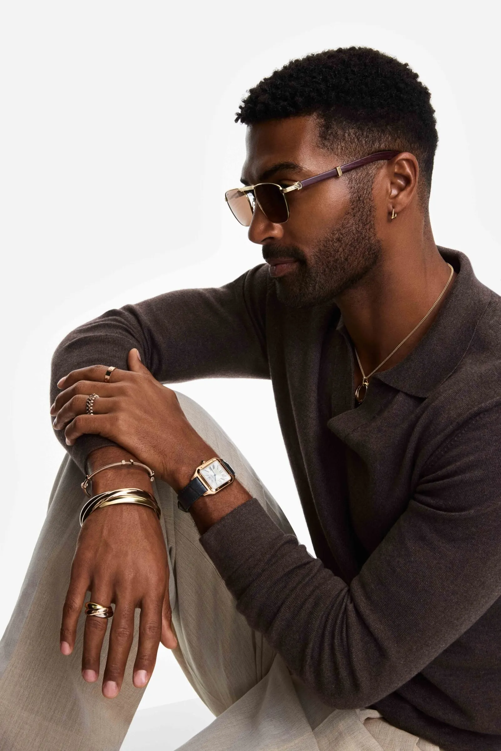Man in brown knit polo and tinted sunglasses wearing gold bracelets, rings and a square watch, seated on white, for Cartier.