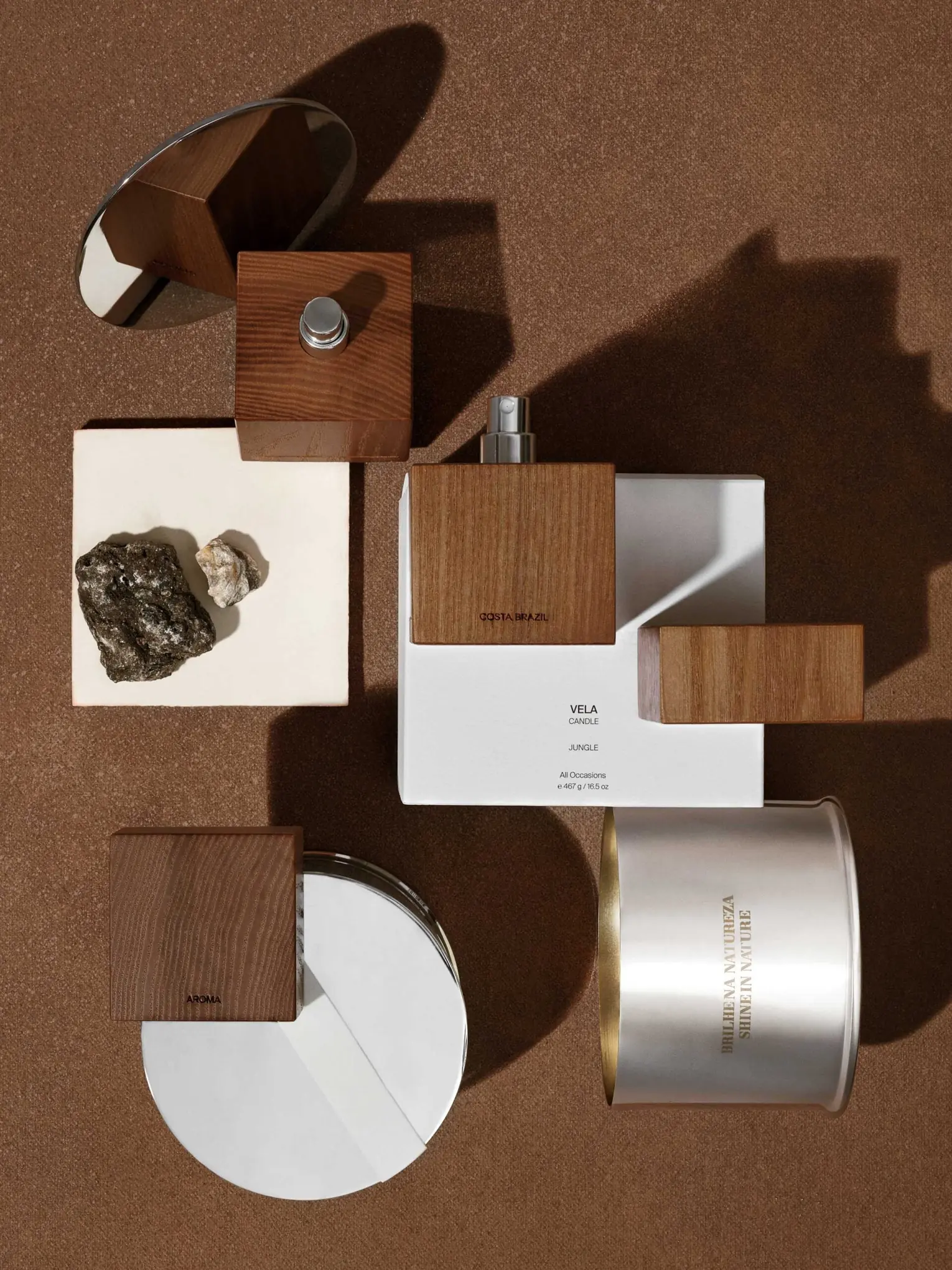 Still life of wood-capped fragrance bottles, Vela candle box, silver tin and stones on brown ground, for Costa Brazil.