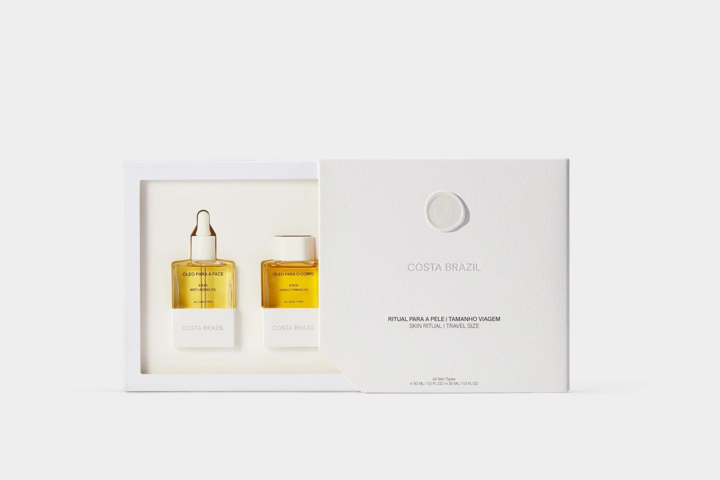 Still life of a white gift box opened to show face and body oil bottles in golden oil on a grey backdrop, for Costa Brazil.