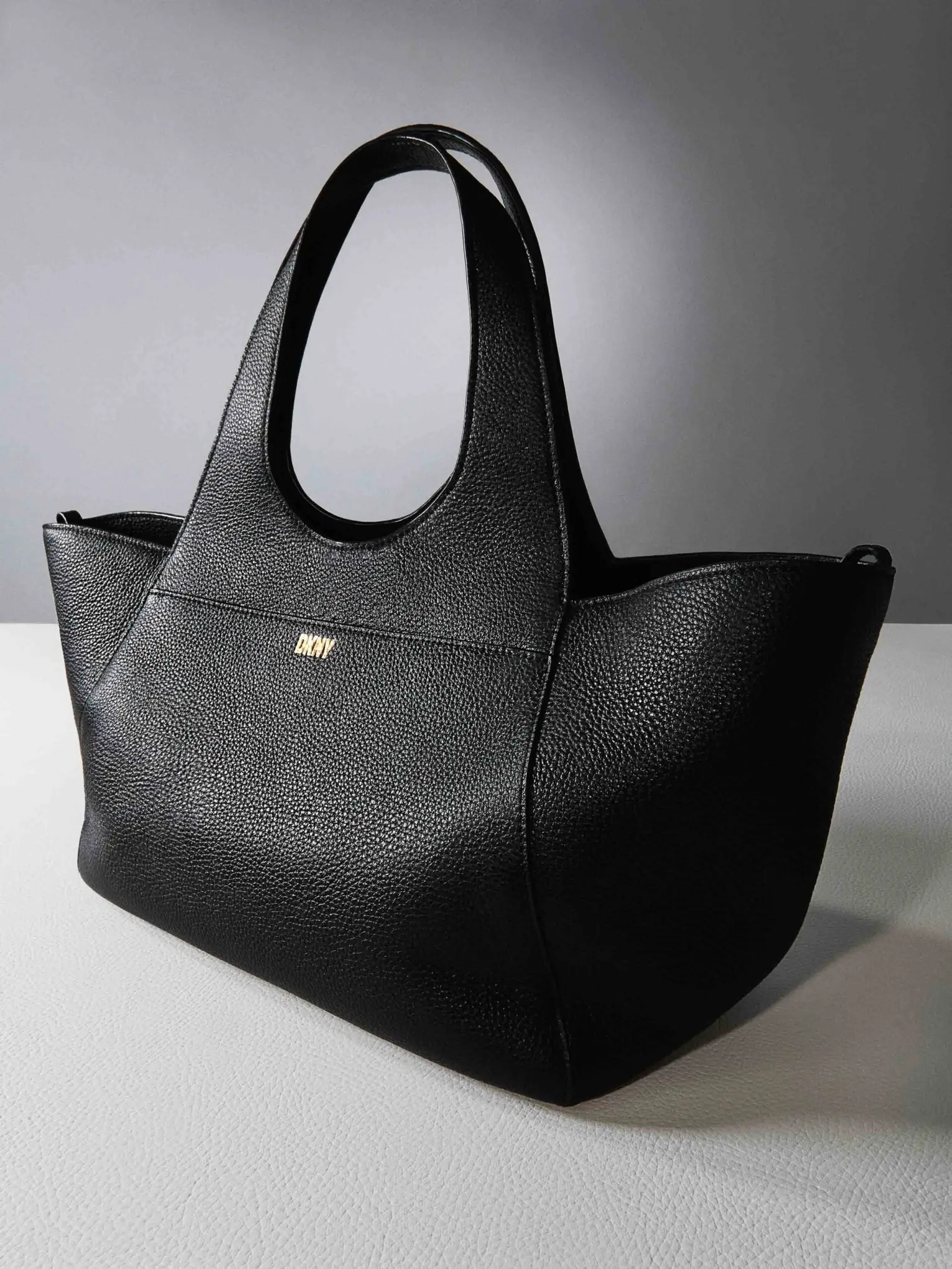 Black pebbled leather tote bag with gold logo on a white surface against a grey backdrop, for DKNY.