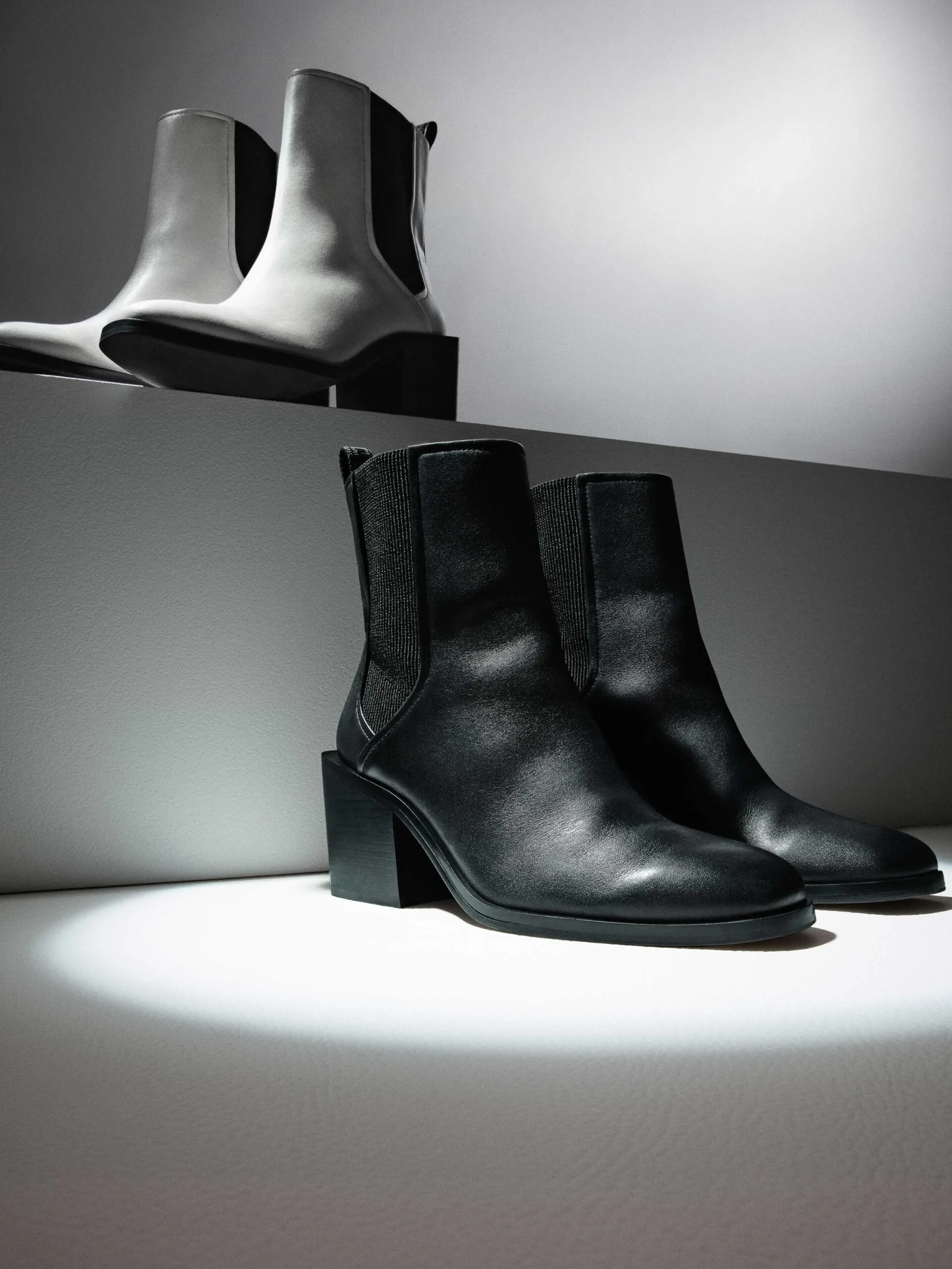 Black leather Chelsea boots in a spotlight below a white pair on a grey step, still life, for DKNY.