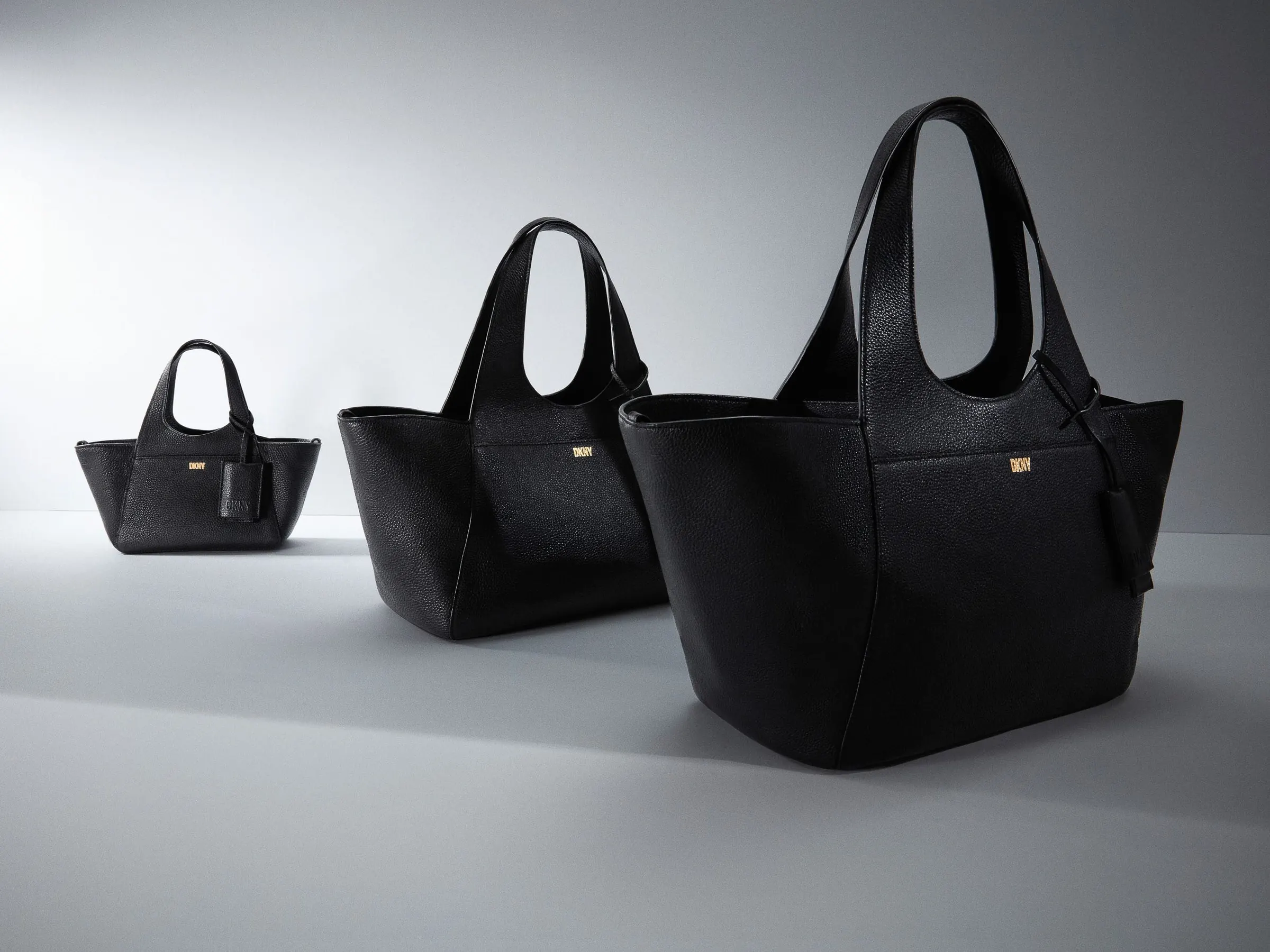 Three black pebbled leather tote bags in graduated sizes with gold logo plates on a grey studio floor, for DKNY.
