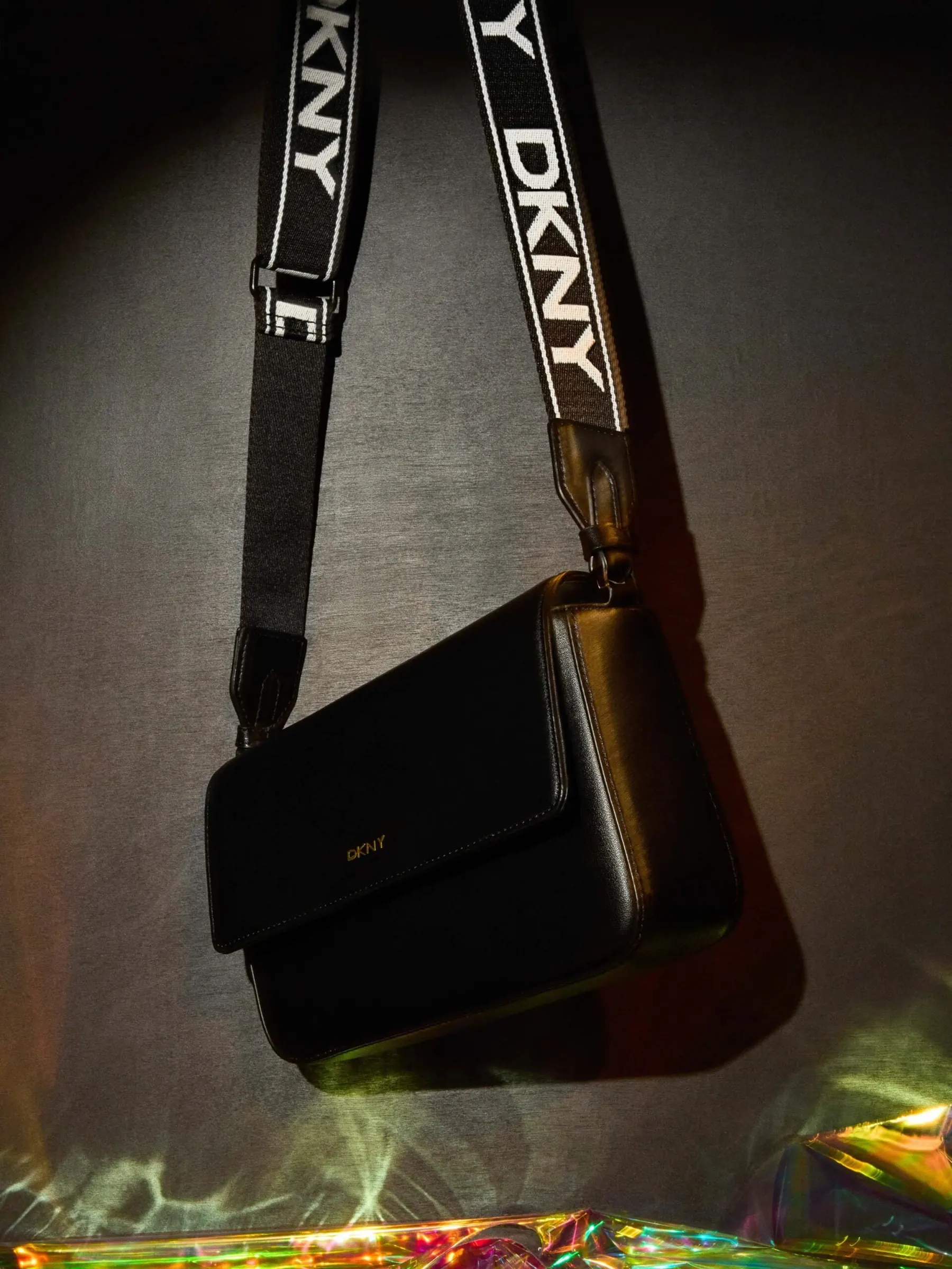 Black leather crossbody bag on a black and white DKNY logo strap, lit over iridescent film on a grey backdrop, for DKNY.