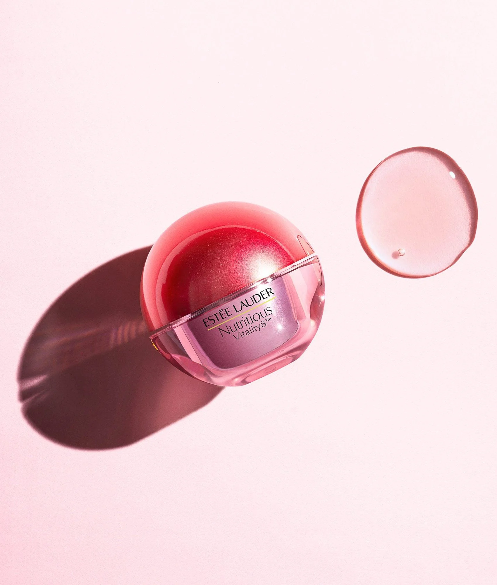 Still life of a red spherical Nutritious Vitality8 jar beside a clear gel swatch on a pale pink surface, for Estée Lauder.