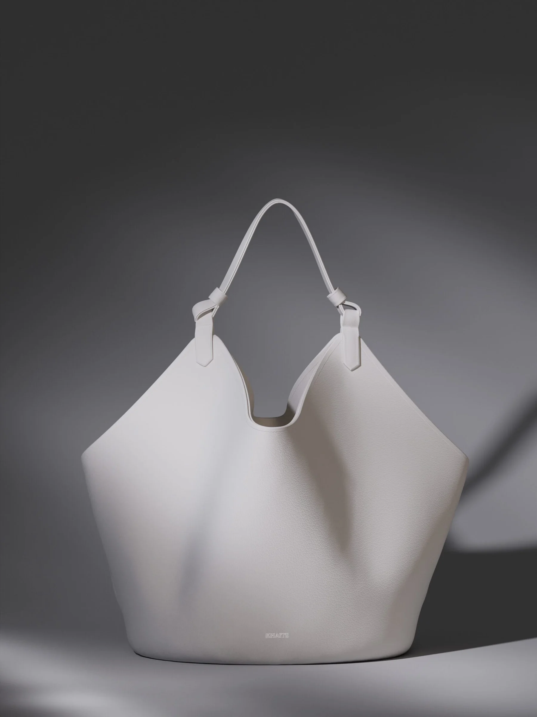 White leather tote bag with knotted shoulder strap under soft shadowed light on a grey backdrop, for Khaite.