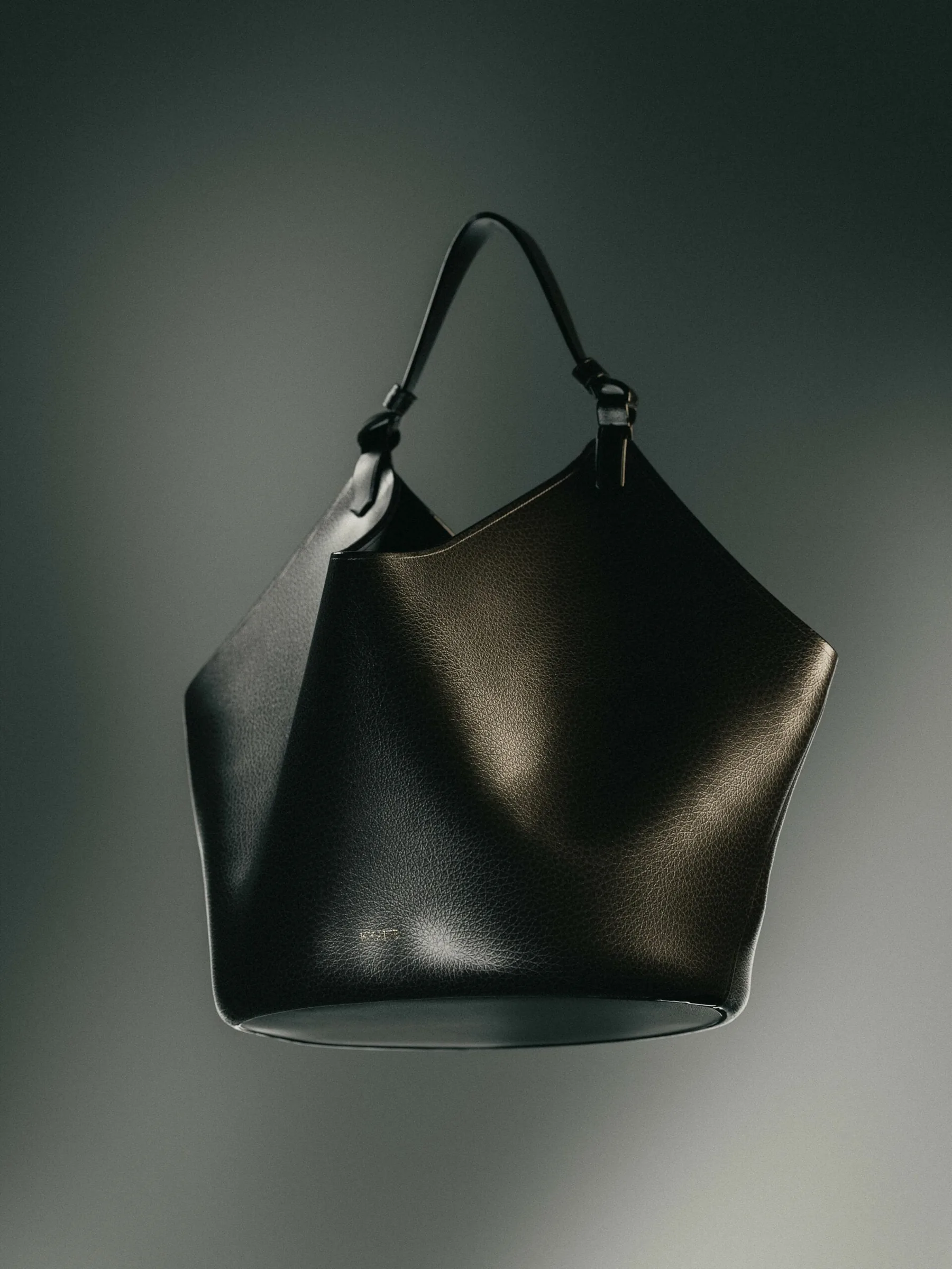 Black grained leather bucket bag with a single top handle, lit softly against a grey backdrop, for Khaite.