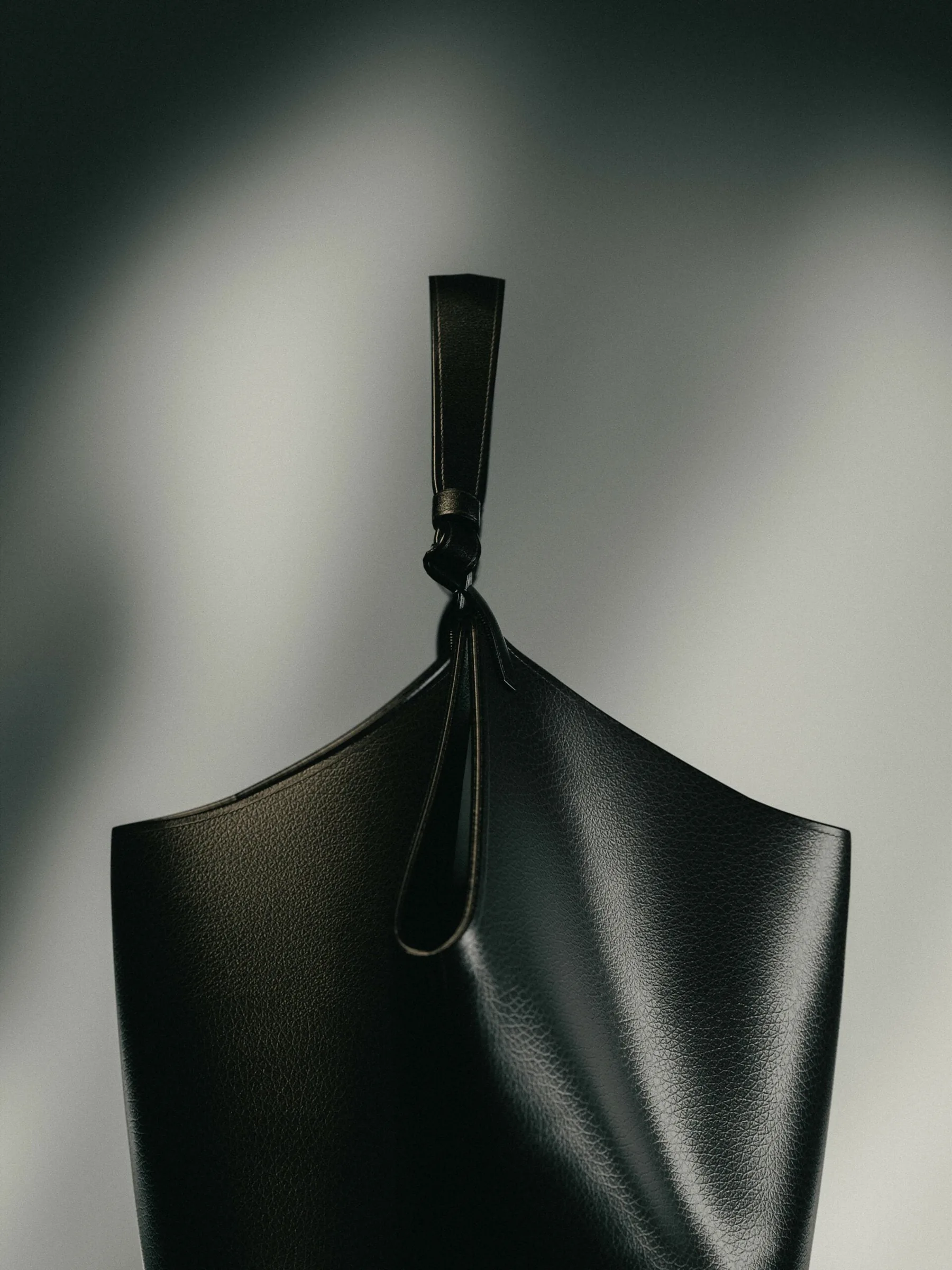 Black grained leather tote with a knotted strap, lit against a grey wall with soft shadow, for Khaite.