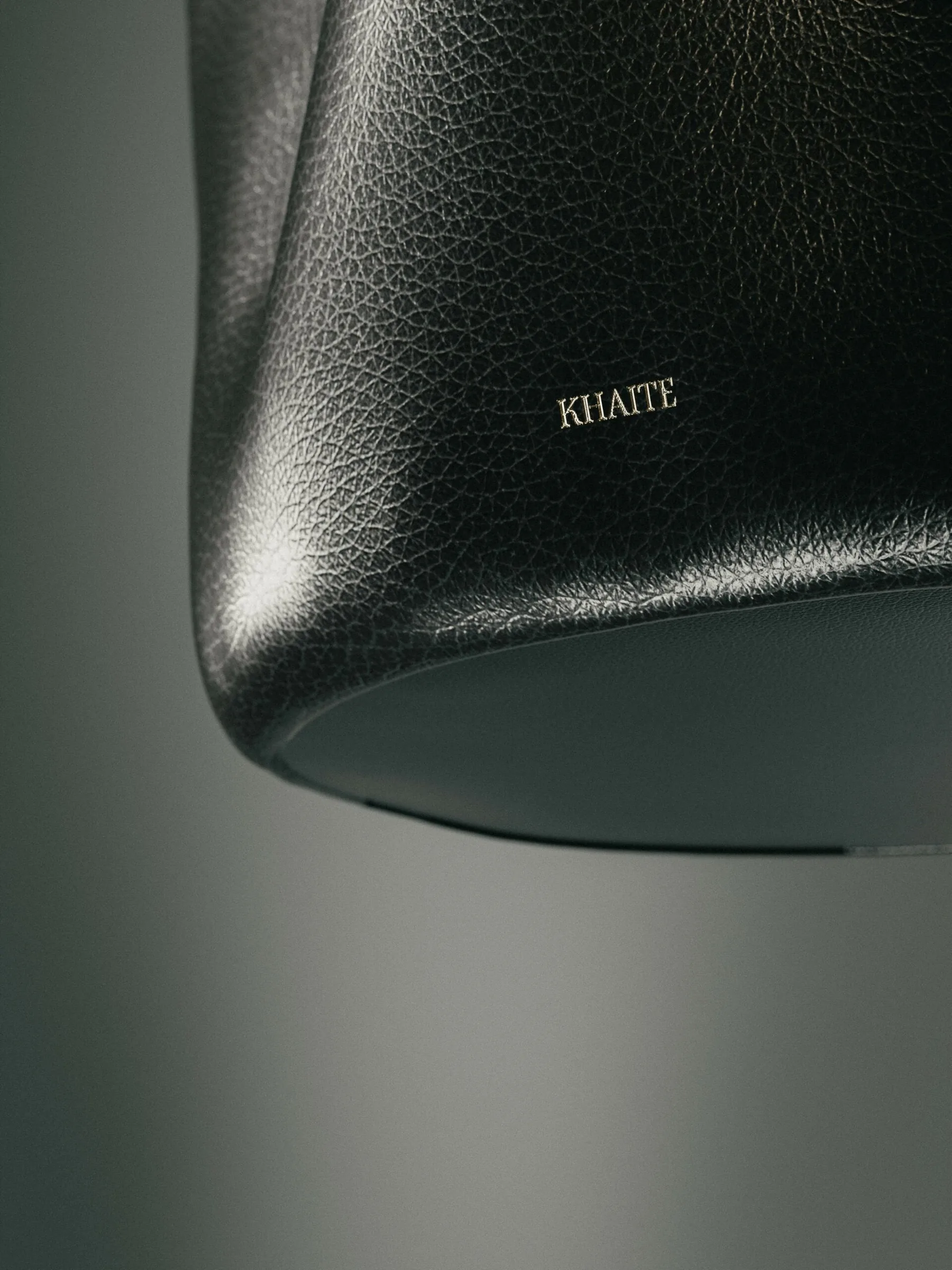 Close-up of a black pebbled leather bag with a gold-stamped logo against a soft grey backdrop, for Khaite.