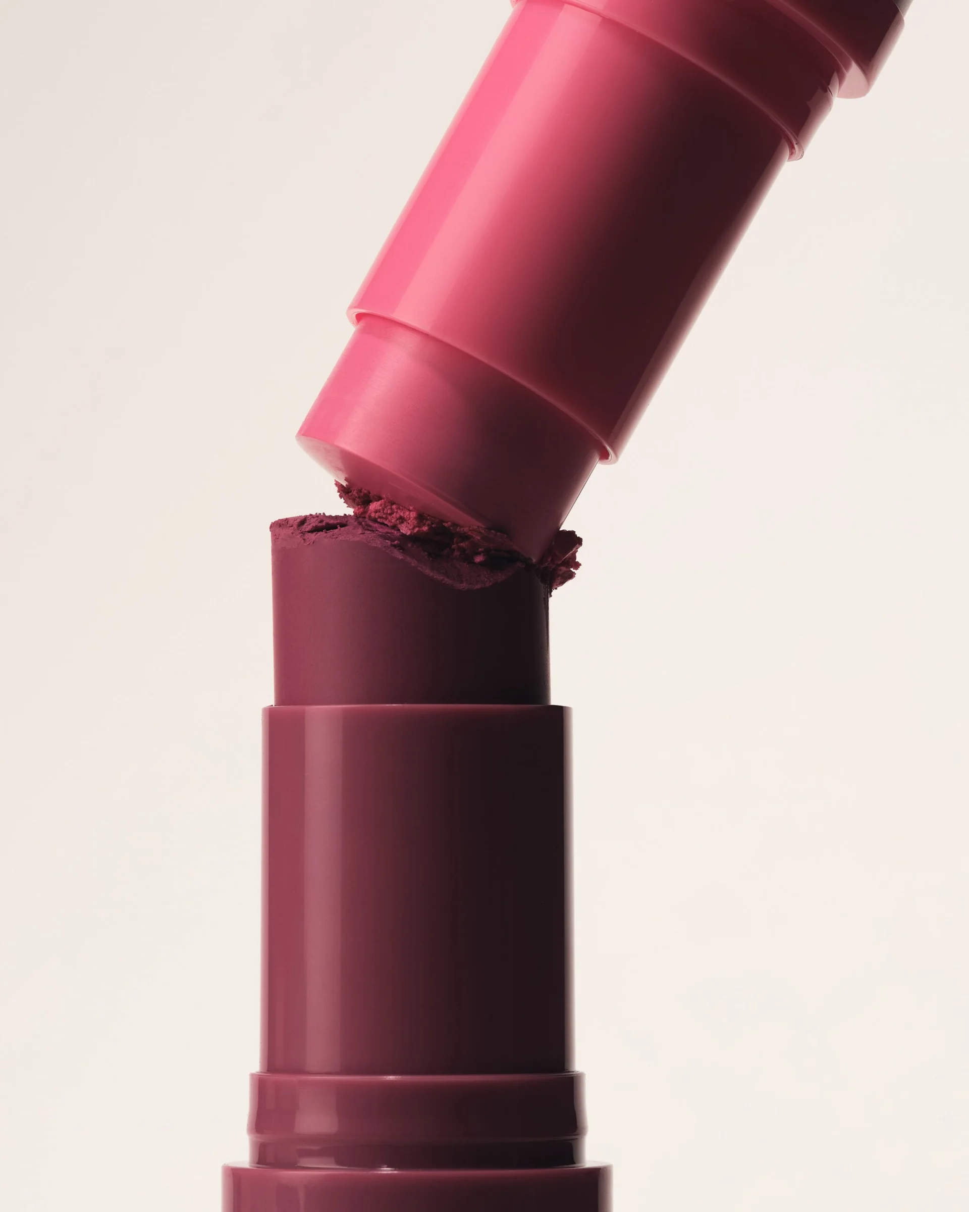 Still life of a pink lipstick tube pressing into a crumbling plum lipstick bullet on a cream background, for Kylie Cosmetics.