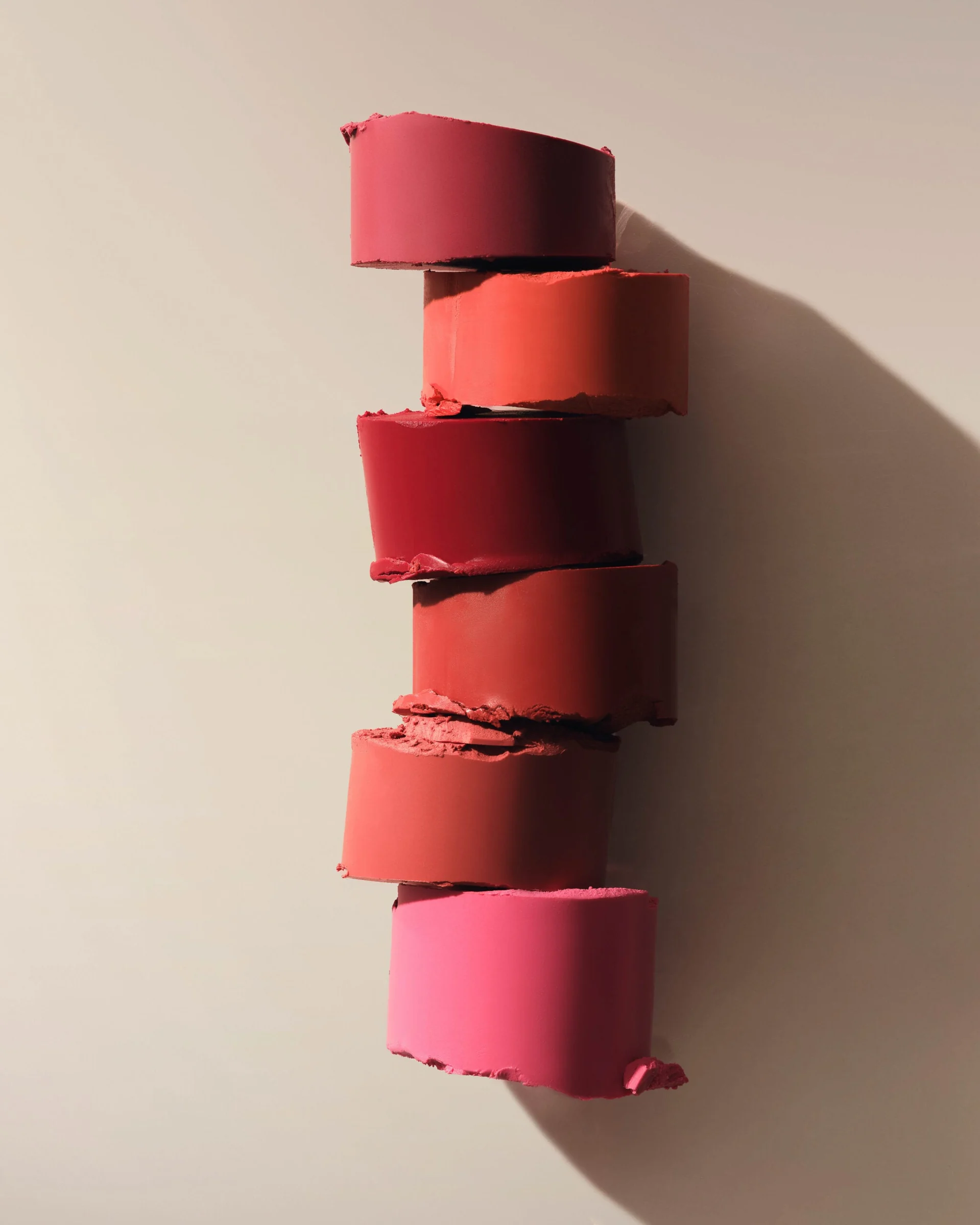 Still life of six lipstick slices in pink, red and coral shades stacked on a beige wall with a long shadow, for Kylie Cosmetics.