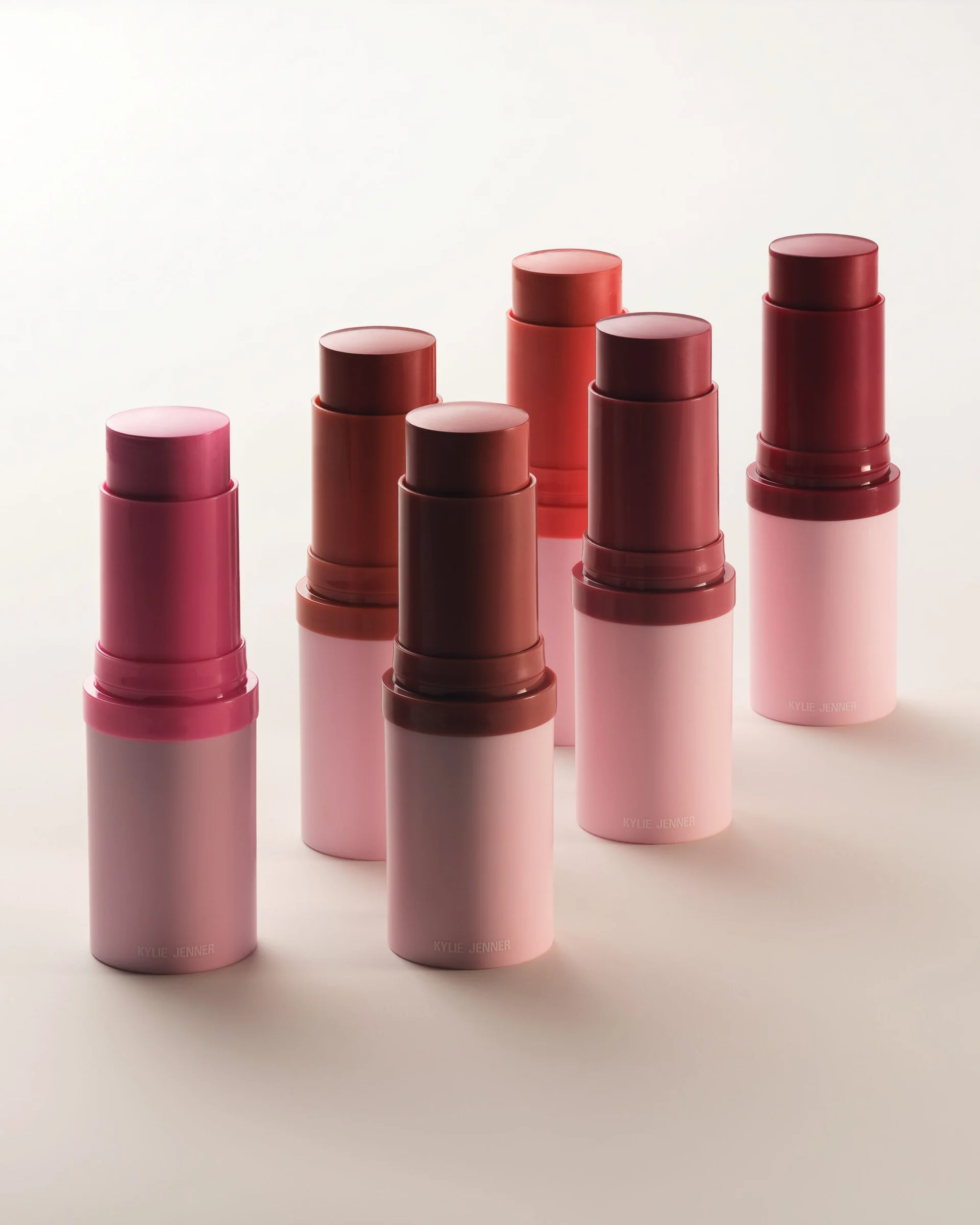 Six cream blush sticks in pink, berry and brick red shades with pale pink bases on a cream backdrop, for Kylie Cosmetics.
