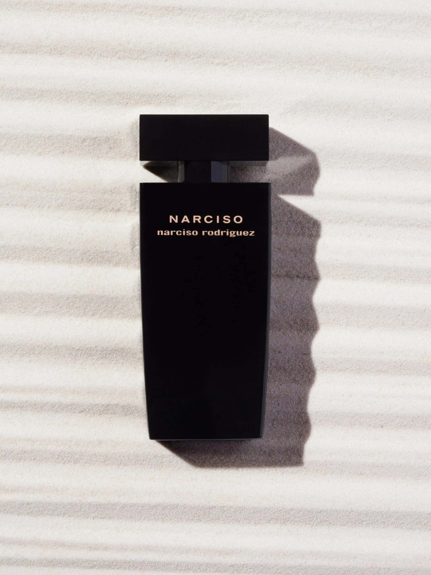 Black NARCISO fragrance bottle lying on rippled white sand under hard sunlight with a long shadow, for Narciso Rodriguez.